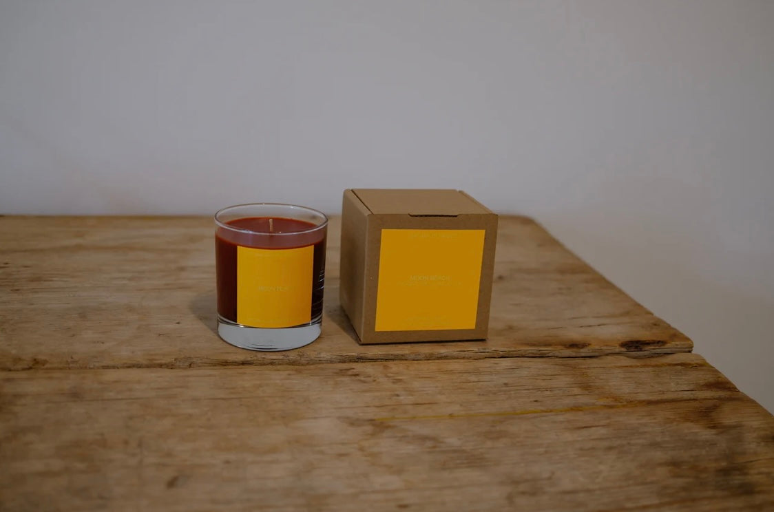 Candle in a glass jar with a yellow label next to its cardboard box on a wooden surface.
