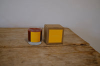 Candle in a glass jar with a yellow label next to its cardboard box on a wooden surface.