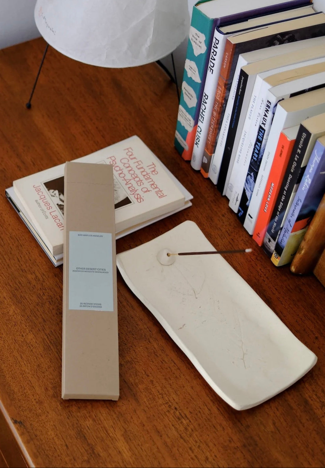Box of incense with a label and an incense stick in a holder on a wooden surface next to books and a white lamp.
