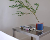 Glass jar candle with a blue label on a side table with a book and plant