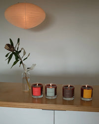Four candles in different colors on a wooden surface with a lamp in the background.