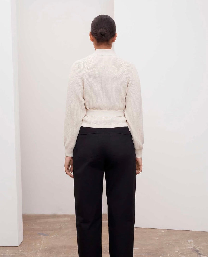 Person wearing a beige sweater and black pants against a white wall.