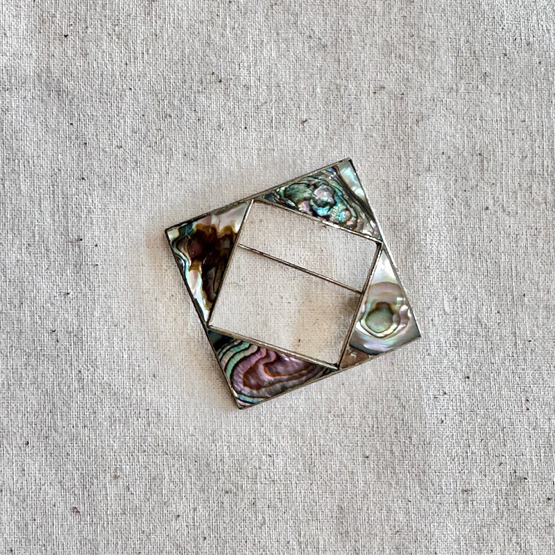 Square decorative brooch with abalone shell inlay on a textured beige background