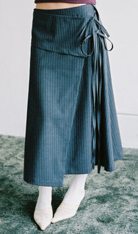 A person wearing a dark grey wrap-style midi skirt with thin vertical pinstripes, a side tie detail, white socks, and white pointed-toe flats, standing on a textured green rug.