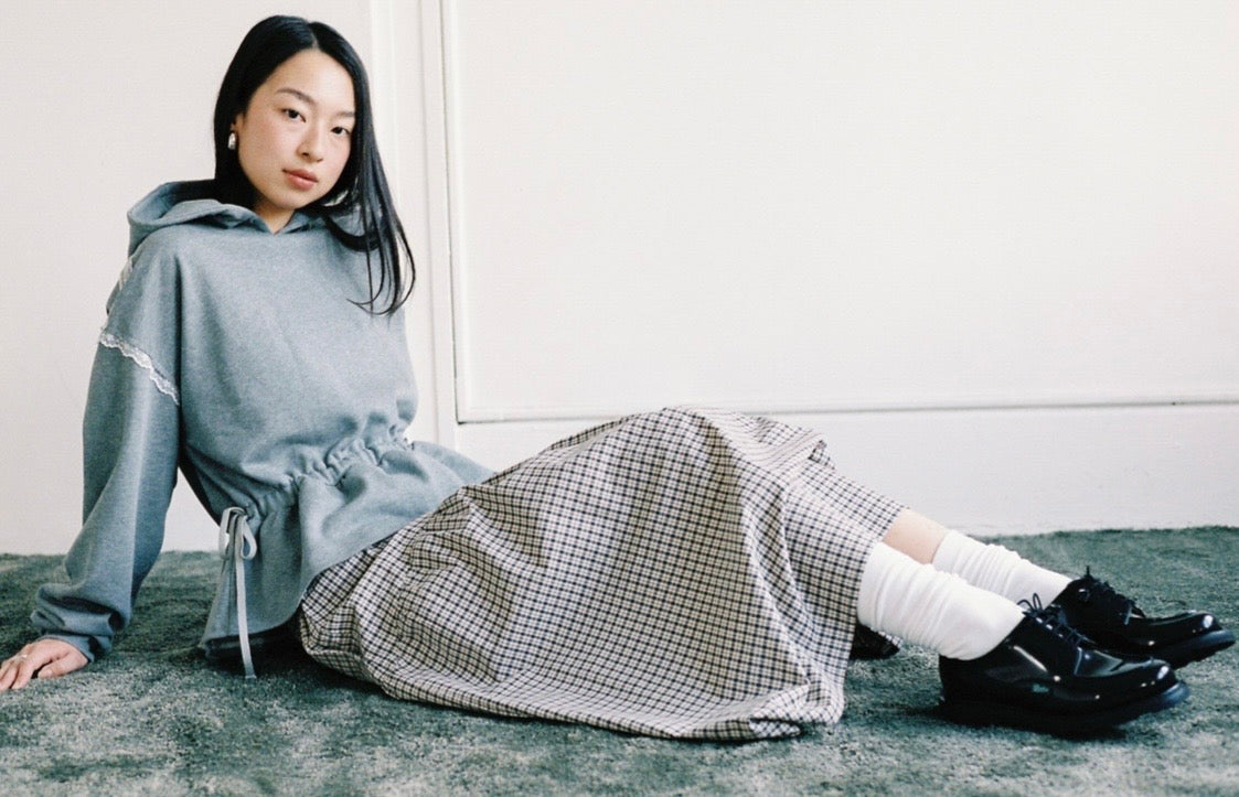 A woman sits on a gray carpet against a white wall, wearing a light gray hooded sweatshirt, a black-and-white gingham skirt, white socks, and black leather shoes.