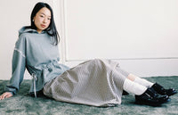 A woman sits on a gray carpet against a white wall, wearing a light gray hooded sweatshirt, a black-and-white gingham skirt, white socks, and black leather shoes.