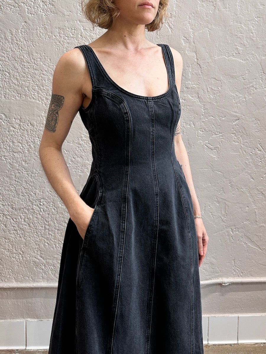 Person wearing a black sleeveless denim dress against a plain wall.