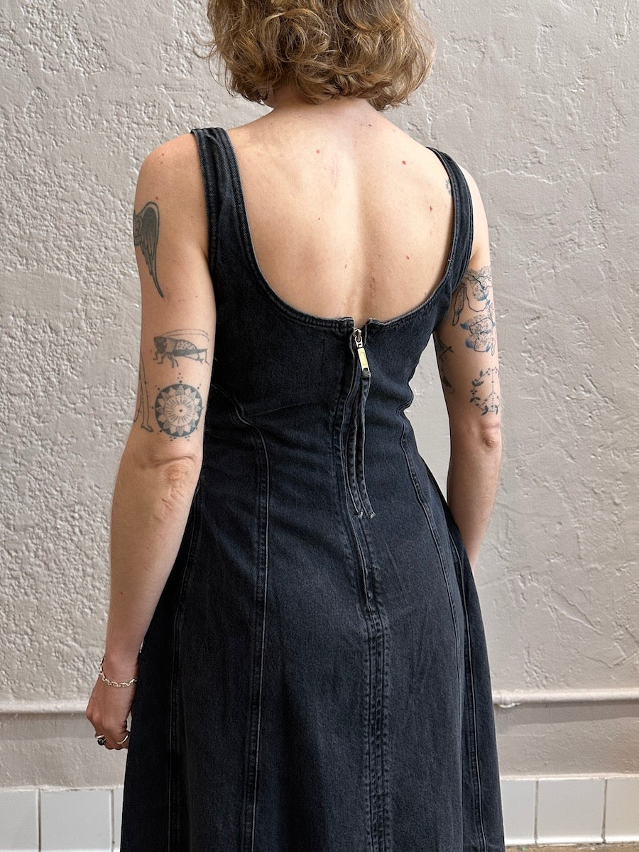 Person wearing a black dress with visible tattoos on arms against a plain wall.