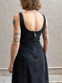 Person wearing a black dress with visible tattoos on arms against a plain wall.