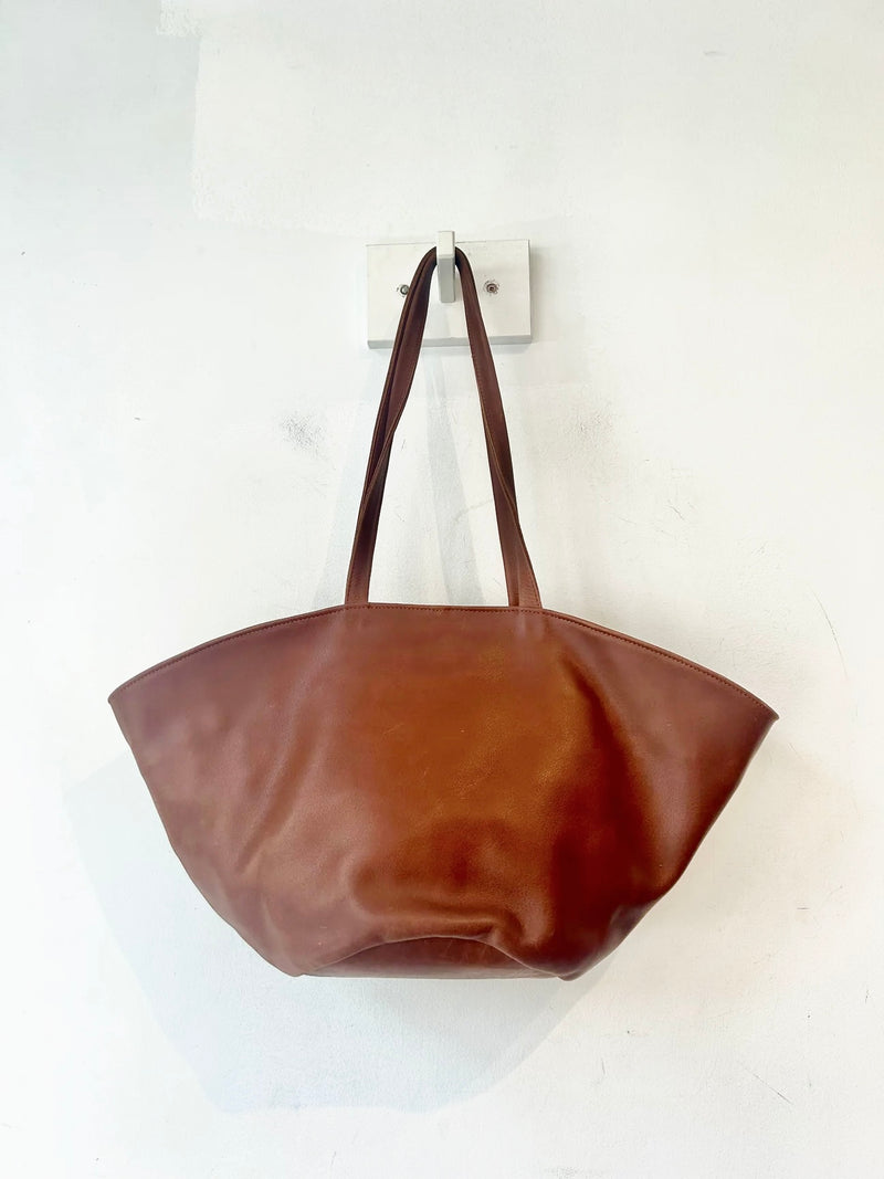 A brown leather tote bag with long shoulder straps, hanging from a wall hook against a plain white wall.