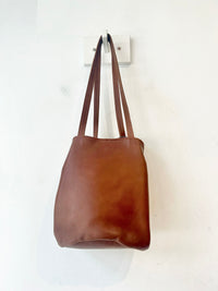 A brown leather shoulder bag with long straps, hanging from a white wall hook against a plain white wall.