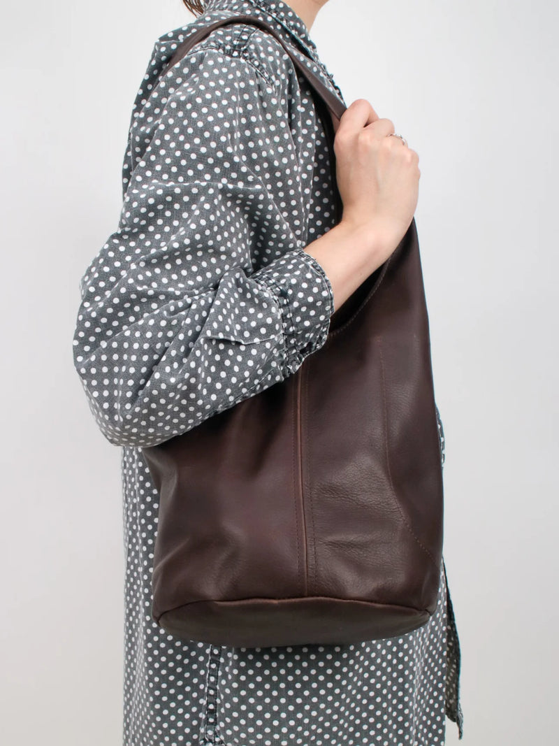 Person with a polka dot shirt wearing a dark brown leather bag over one shoulder against a plain background