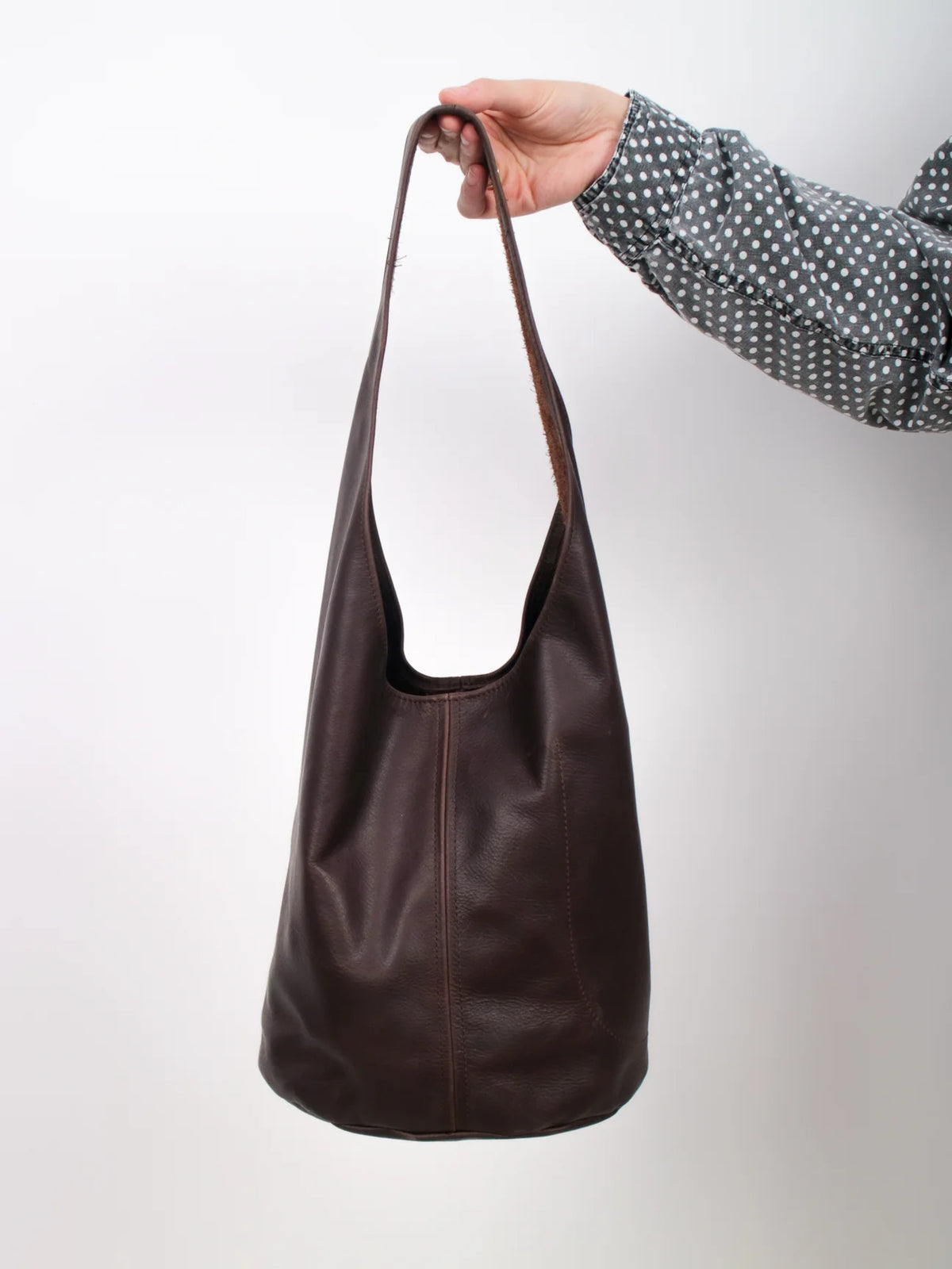 A dark brown leather slouchy shoulder bag held by a hand wearing a black and white polka dot shirt, set against a plain light gray background.