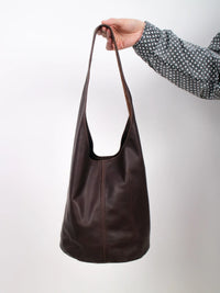 A dark brown leather slouchy shoulder bag held by a hand wearing a black and white polka dot shirt, set against a plain light gray background.