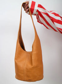 A tan leather hobo shoulder bag held by a hand wearing a red and white striped shirt, set against a plain light gray background.