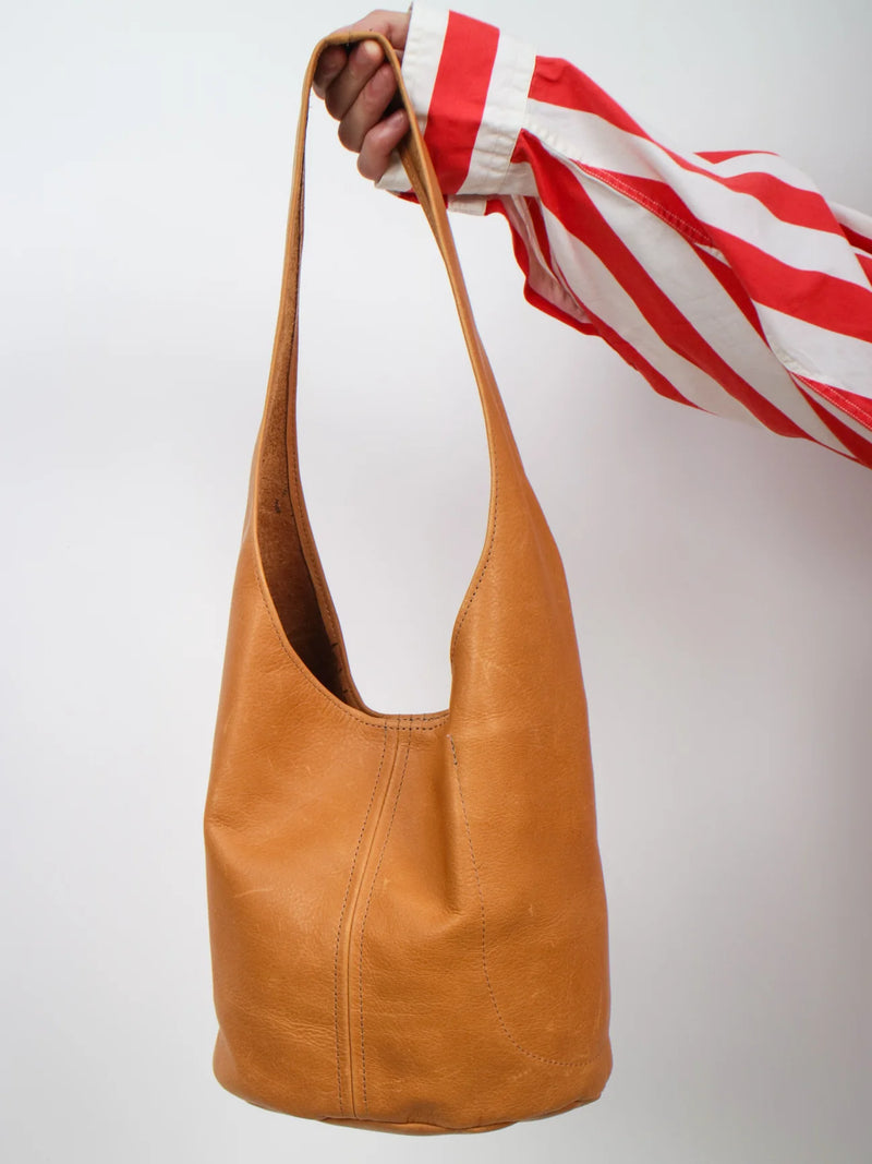 A tan leather hobo shoulder bag held by a hand wearing a red and white striped shirt, set against a plain light gray background.