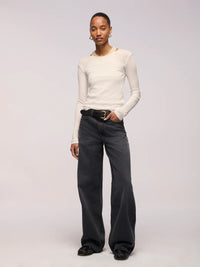 A model stands against a light gray studio background wearing a fitted off-white long-sleeve top, dark charcoal wide-leg jeans with a black belt, and black shoes.