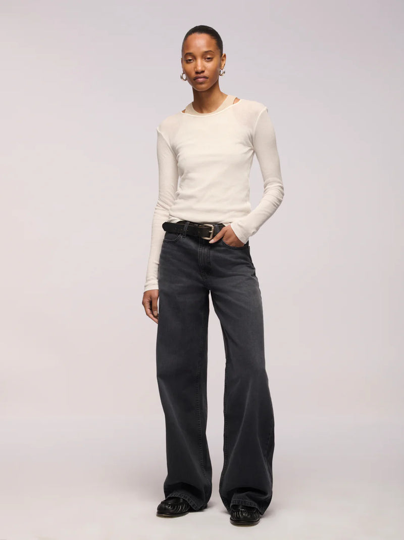A model stands against a light gray studio background wearing a fitted off-white long-sleeve top, dark charcoal wide-leg jeans with a black belt, and black shoes.