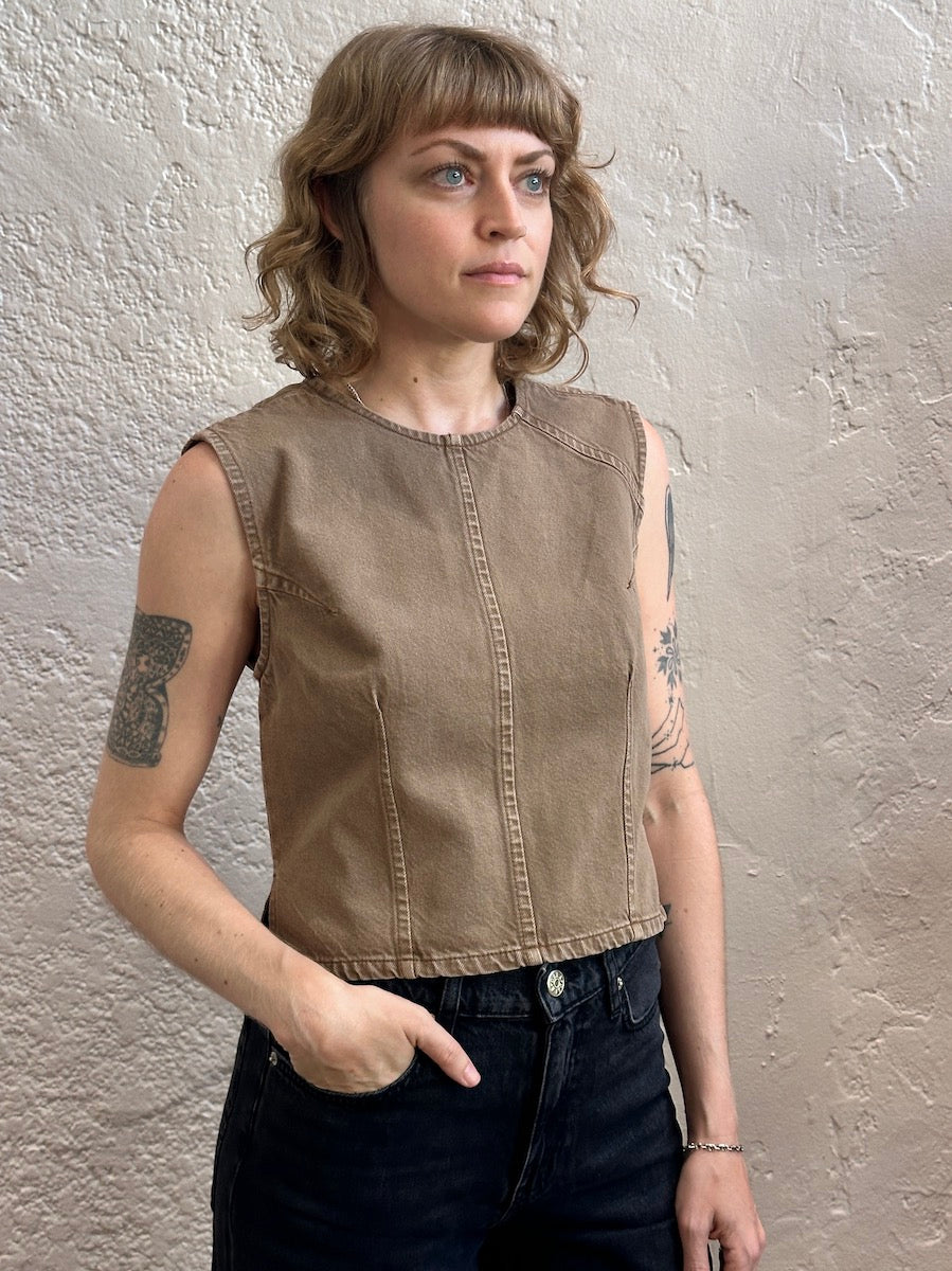 A woman with shoulder-length blonde hair and tattoos wearing a sleeveless, cropped tan-brown denim top over dark jeans, standing against a textured beige wall.
