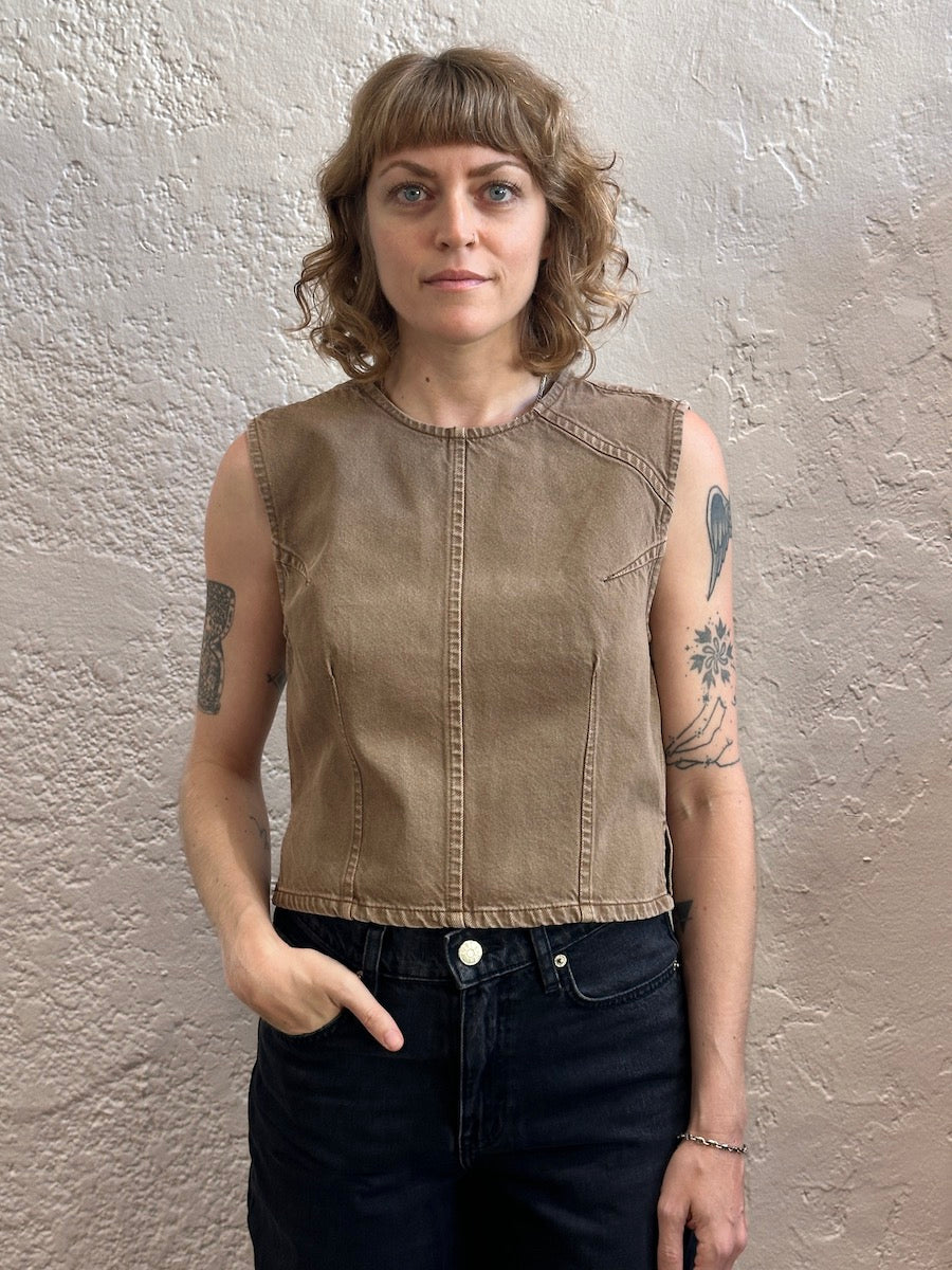 A woman with shoulder-length wavy blonde hair stands against a textured beige plaster wall, wearing a sleeveless cropped denim top in a warm taupe-brown color and dark jeans.