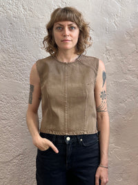 A woman with shoulder-length wavy blonde hair stands against a textured beige plaster wall, wearing a sleeveless cropped denim top in a warm taupe-brown color and dark jeans.
