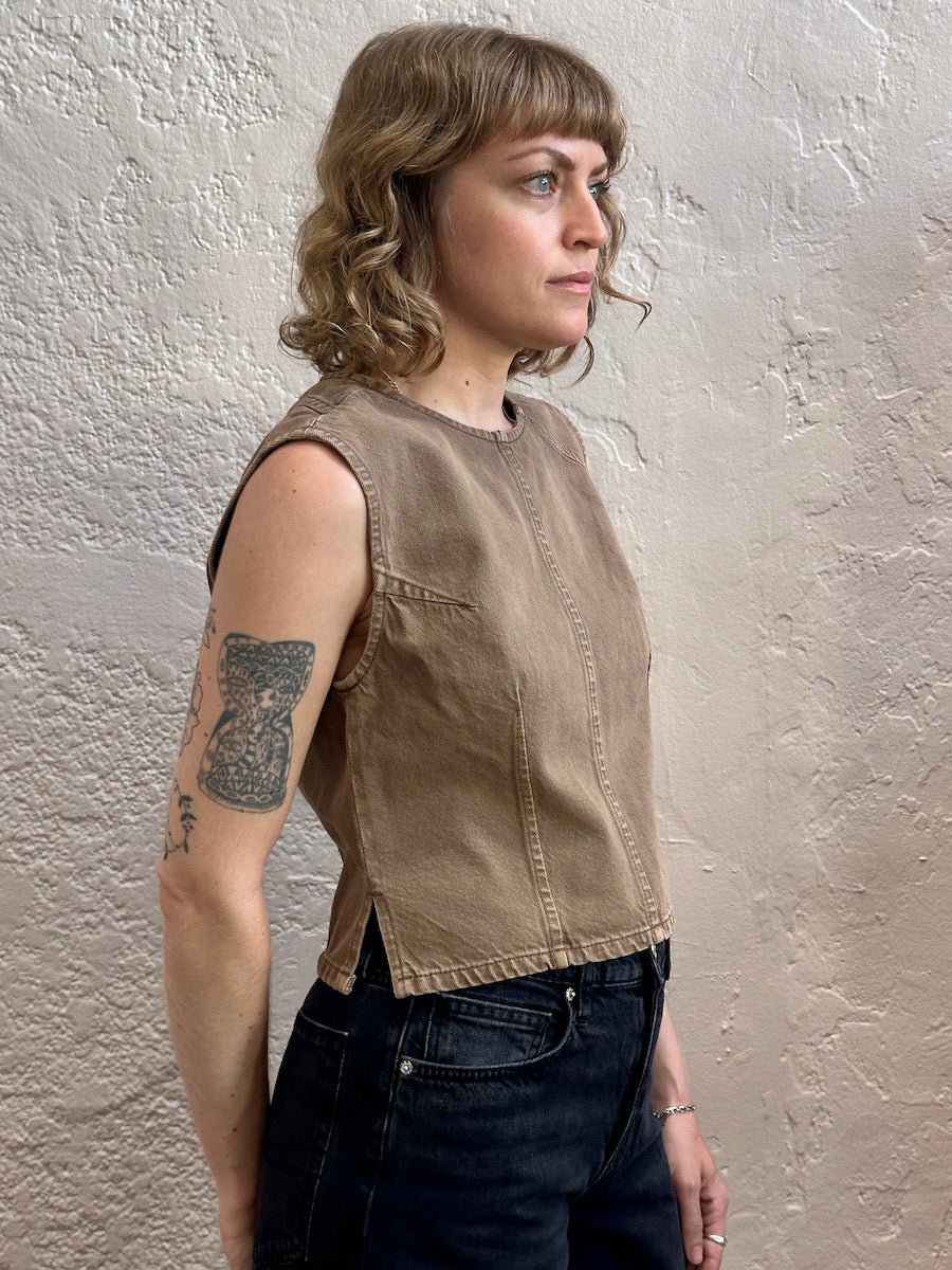A woman with shoulder-length blonde hair and a tattoo on her arm is shown in profile wearing a sleeveless, cropped tan utility-style top and dark jeans, standing against a textured beige plaster wall.