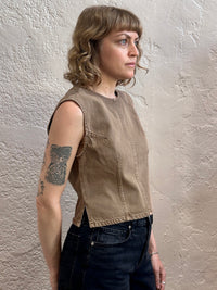 A woman with shoulder-length blonde hair and a tattoo on her arm is shown in profile wearing a sleeveless, cropped tan utility-style top and dark jeans, standing against a textured beige plaster wall.