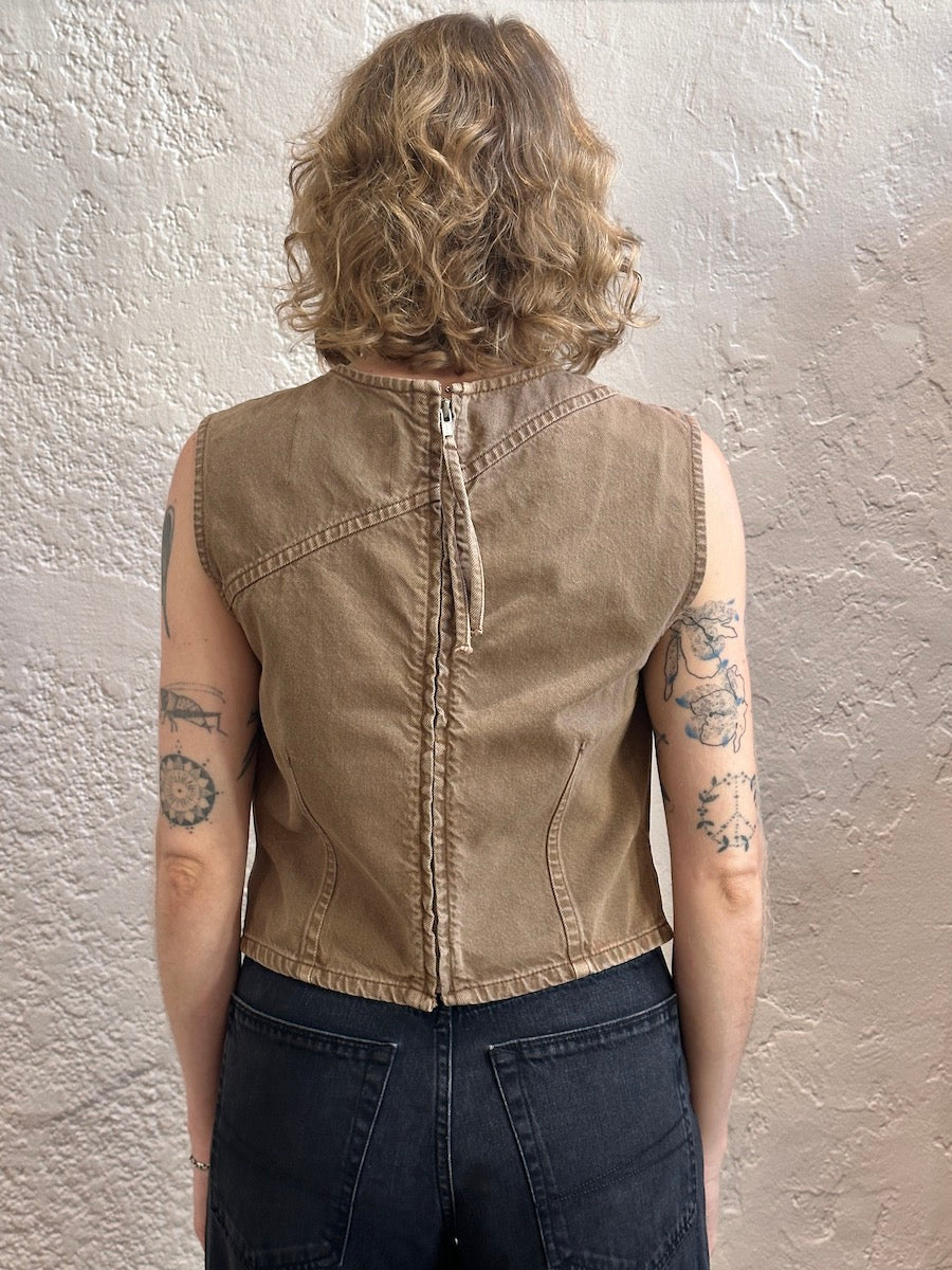 A person shown from the back wearing a sleeveless cropped tan sleeveless top with a central zipper and dark jeans, standing against a textured off-white wall.