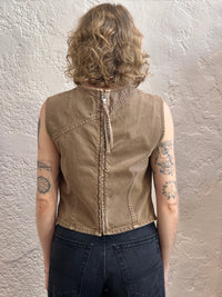 A person shown from the back wearing a sleeveless cropped tan sleeveless top with a central zipper and dark jeans, standing against a textured off-white wall.