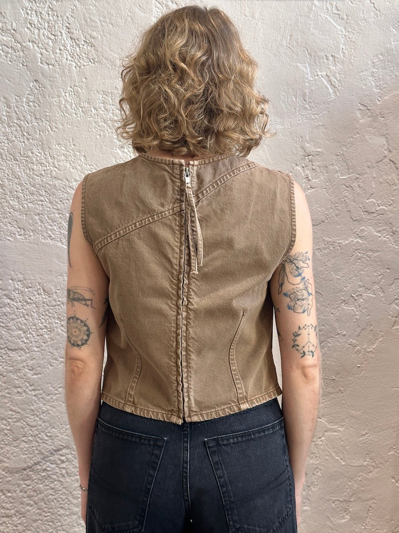 A person shown from the back wearing a sleeveless cropped tan sleeveless top with a central zipper and dark jeans, standing against a textured off-white wall.