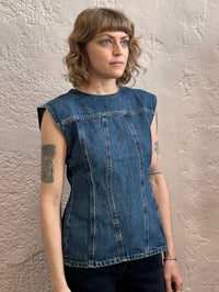 A woman with shoulder-length blonde hair and tattoos wearing a sleeveless blue denim top, standing against a textured beige stucco wall.