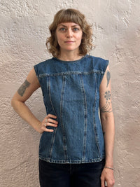 A woman with shoulder-length blonde hair and tattoos wearing a sleeveless blue denim top, standing against a textured beige plaster wall.