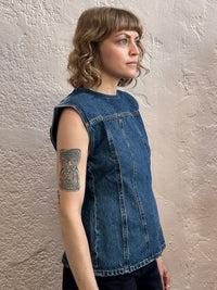 A woman with shoulder-length blonde hair and a side tattoo is wearing a sleeveless blue denim top, standing against a textured beige plaster wall.