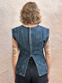 A woman viewed from behind wearing a sleeveless blue denim vest with a central zipper and gathered seams, standing against a textured light beige wall.