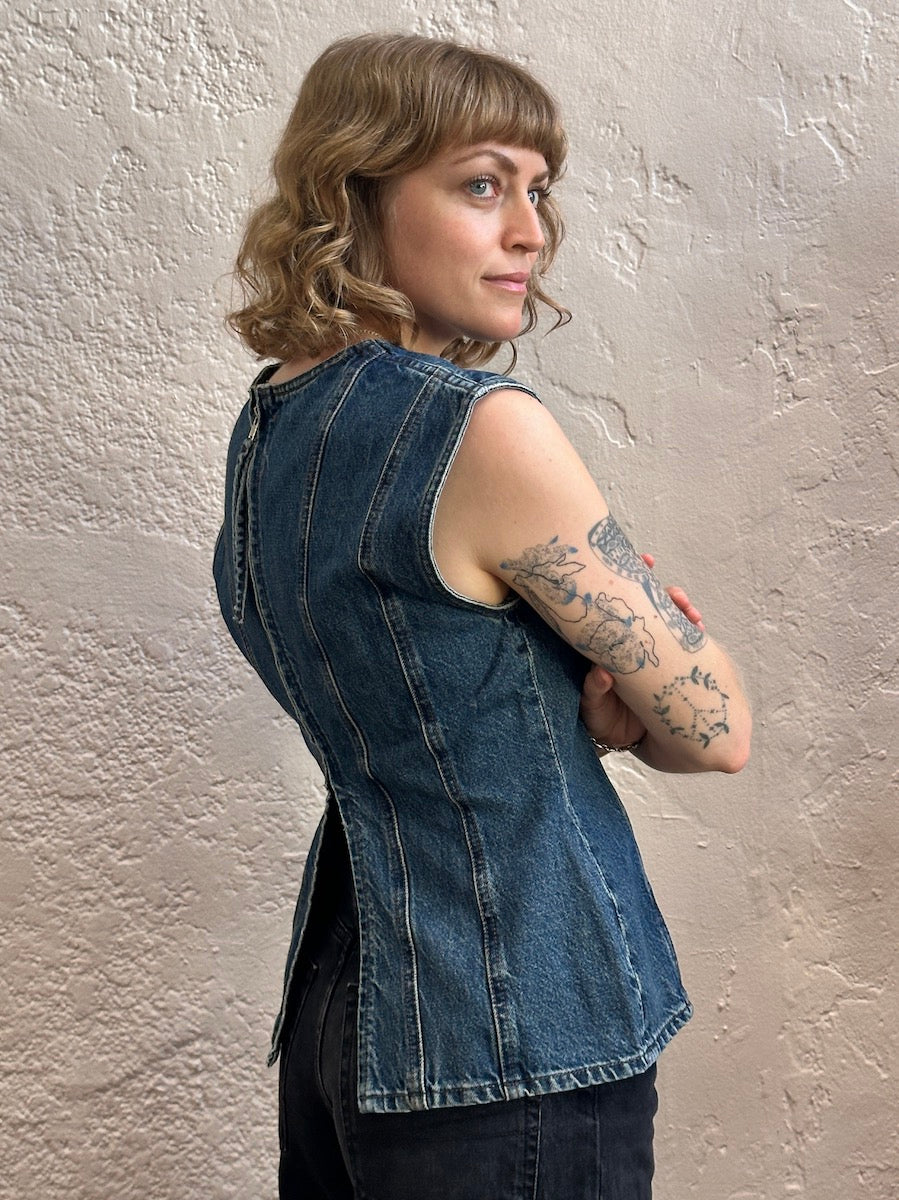 A woman with shoulder-length blonde wavy hair and tattoos on her arm, wearing a sleeveless dark blue denim vest, posing against a textured beige plaster wall.