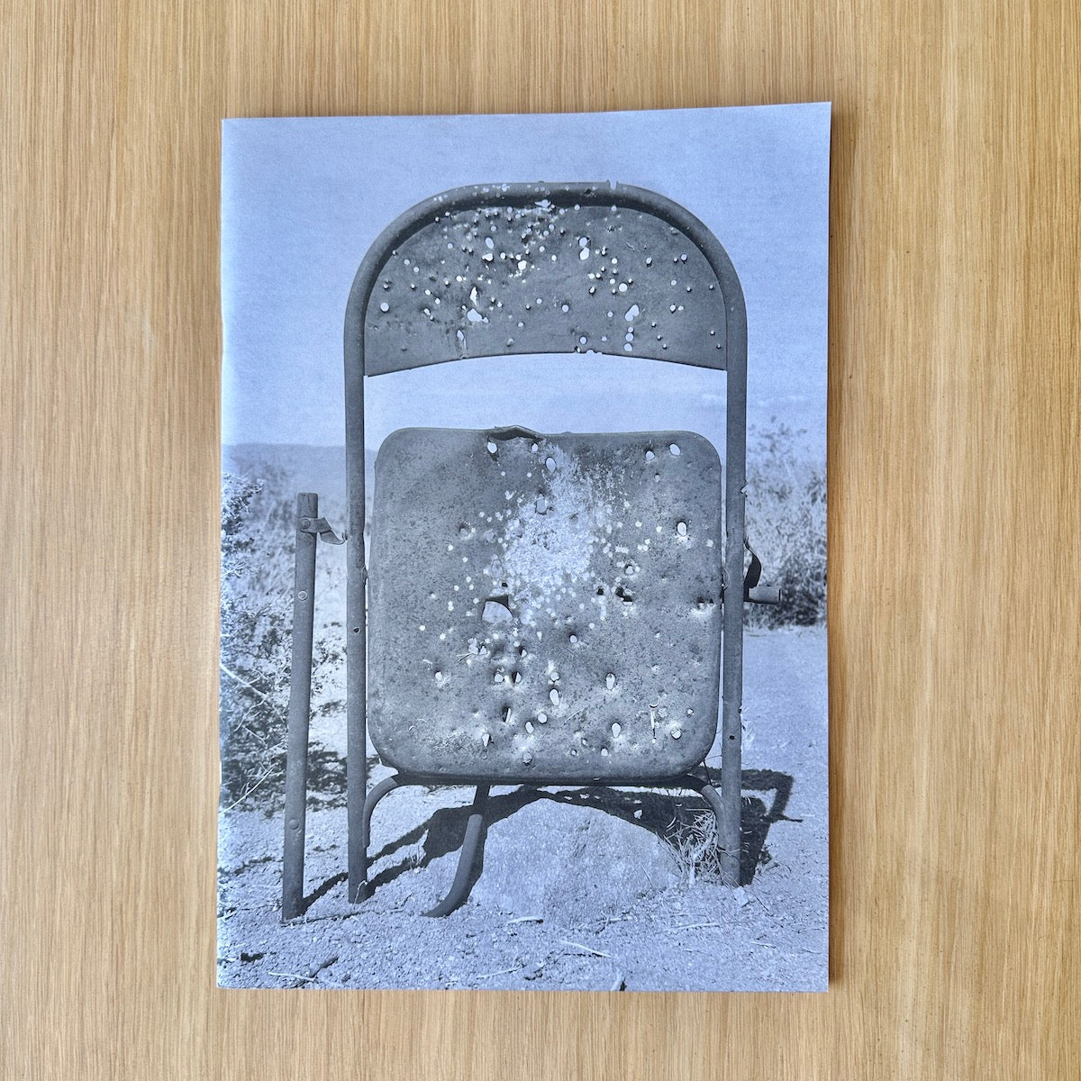 A print publication featuring a heavily bullet-riddled folding chair in a desert landscape, displayed flat on a light wood grain surface.