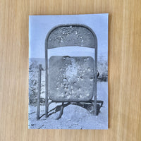 A print publication featuring a heavily bullet-riddled folding chair in a desert landscape, displayed flat on a light wood grain surface.