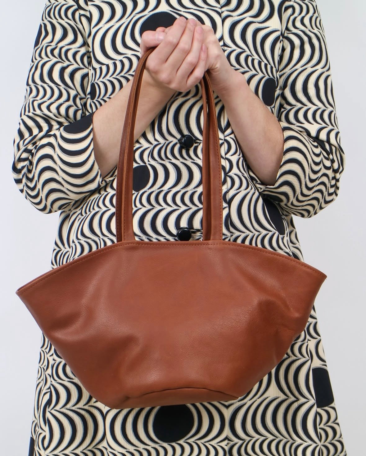 A person wearing a black and beige wavy-patterned dress holds a brown leather tote bag against a plain light gray background.