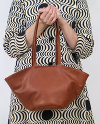 A person wearing a black and beige wavy-patterned dress holds a brown leather tote bag against a plain light gray background.