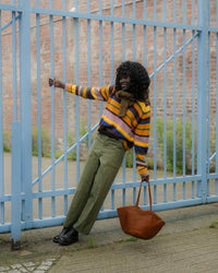 A person stands outdoors in front of a blue metal gate and brick wall, wearing a colorful striped sweater, olive green trousers, black shoes, and holding a brown tote bag.