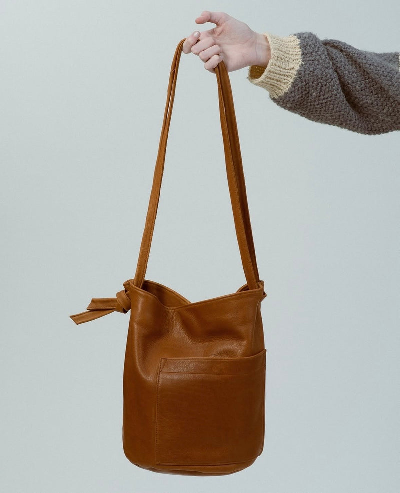 A medium brown leather bucket bag with a shoulder strap, held by a person's hand against a light gray background.