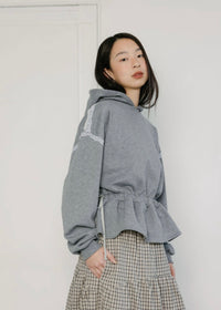 Woman wearing a gray hoodie with cinched waist and a plaid skirt with pockets on a white background
