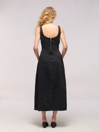 A woman viewed from the back wearing a sleeveless black midi dress with a deep neckline and a central zipper, paired with black heels, against a light gray studio background.