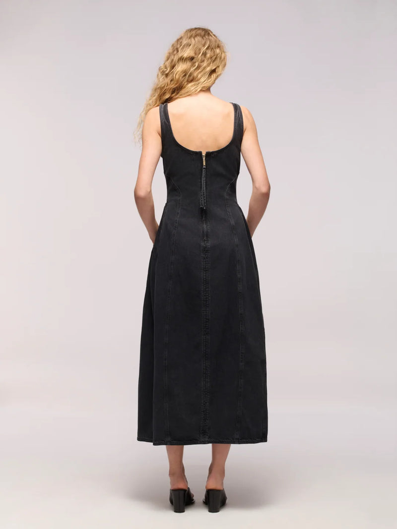A woman viewed from the back wearing a sleeveless black midi dress with a deep neckline and a central zipper, paired with black heels, against a light gray studio background.