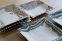 A stack of square ceramic trays with a white base and marbled, fluid nerikomi patterned borders in earthy brown, beige, gray, blue, and green tones, arranged on a light wood surface.