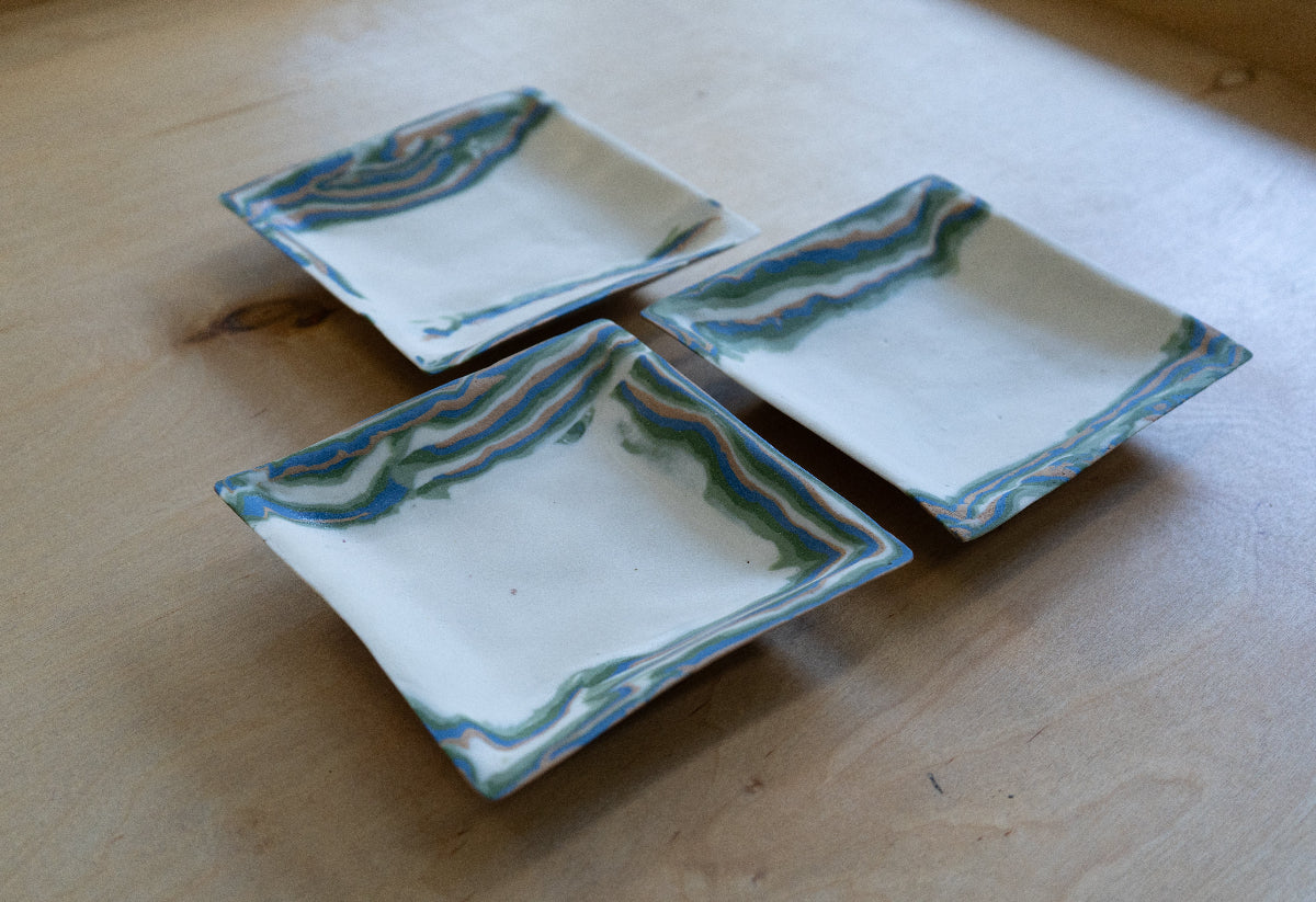 Three square white ceramic trays with wavy blue, green, and orange marbled nerikomi borders, arranged on a light wood tabletop.