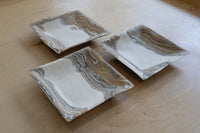 Three square ceramic trays with a white base and swirling brown, tan, and gold agate-like nerikomi patterns, arranged on a light wood tabletop.