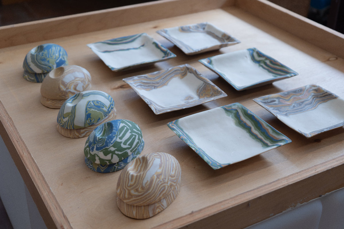 A set of handmade ceramic dishes, including square trays and small pebbles, decorated with marbled nerikomi patterns in blue, green, beige, and white, arranged on a light wood tray.