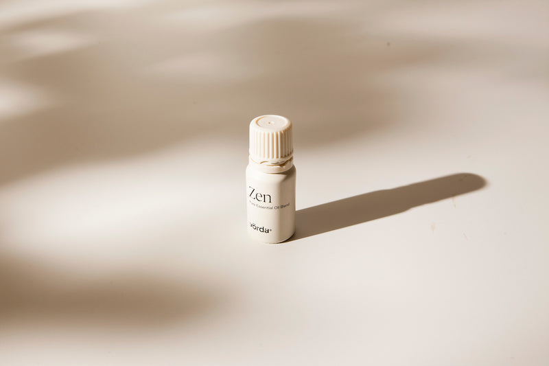A small white bottle labeled 'Zen' and 'vörda' stands on a warm off-white surface with soft shadows cast across it.