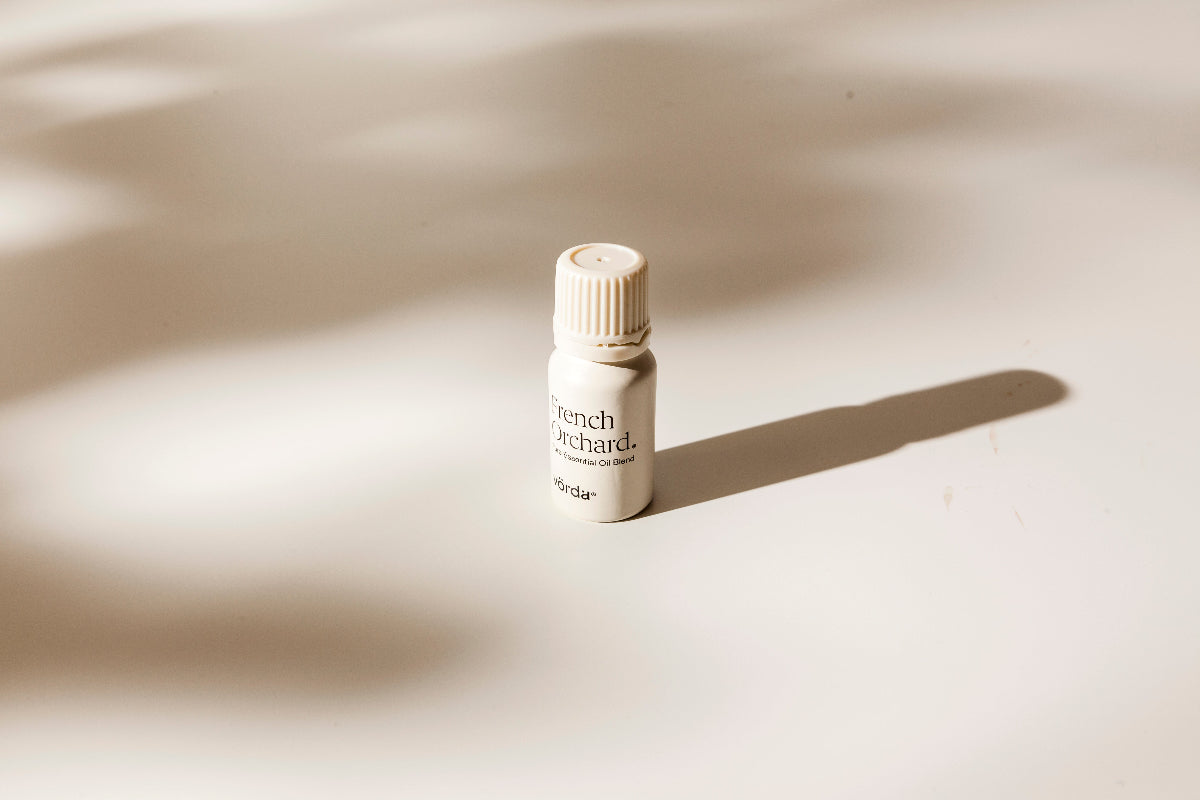 A small cream-colored bottle with a ribbed cap stands on a warm beige surface under soft, directional sunlight that casts a long shadow. The label reads 'French Orchard' and 'vörda'.